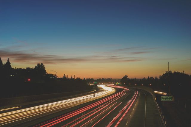 The photo captures a busy highway with car light trails during sunset, creating a sense of motion and speed. The vivid colors in the evening sky contribute to a tranquil yet dynamic urban scene. Ideal for illustrating concepts of transportation, busy city life, and modern infrastructure. It can be used in articles, advertisements, or web designs related to travel, commuting, and city living.