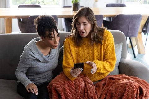 Friends sharing smartphone on sofa, cozy home moment with mustard sweater and rust throw