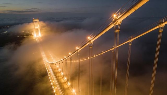 Glowing suspension bridge cutting through fog, illuminated roadway, cables, aerial dusk view