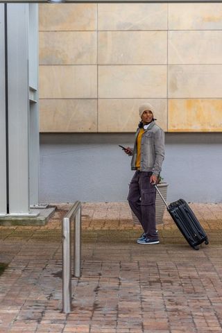 Walking traveler pulling rolling suitcase and checking smartphone in urban plaza