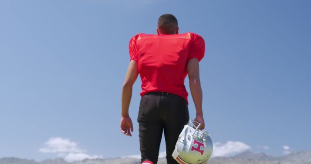 Football player standing with helmet under blue sky is great for themes of sports motivation, team preparation, and fitness marketing. Ideal for use in advertisements, sports articles, and promotional materials. Highlights the determination and professionalism of athletes.