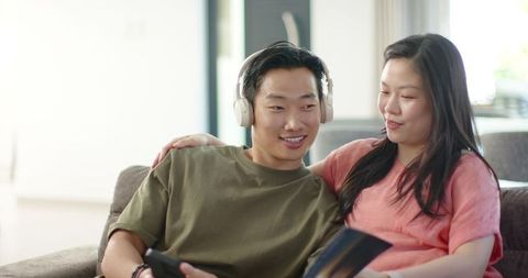 Asian couple relaxing on sofa listening to music and reading in modern living room