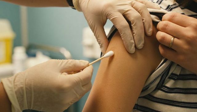 Clinician cleaning upper arm with swab before vaccination, gloved hands, patient in striped shirt