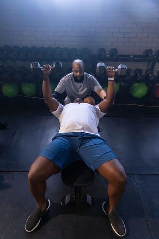 African men performing dumbbell bench press with spotter in gym, trainer guiding technique