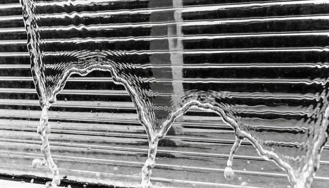 Streaming water flowing over ribbed glass forming arched rivulets and droplets, monochrome