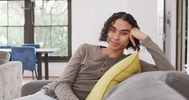 Young man with curly hair is relaxing on a comfortable couch at home. He is smiling and leaning on a yellow pillow. This can be used to depict relaxation, cozy home environments, leisure time, and contentment. Suitable for articles, blogs, or ads focusing on home lifestyle, relaxation products, interior design, or health and well-being.