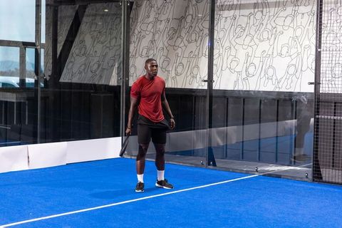 Male athlete standing on blue padel court gripping racket wearing red shirt and black shorts