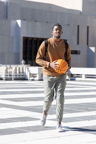 Sunlit African American man walking with basketball in urban plaza carrying backpack