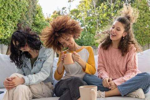 Three diverse women laughing and sipping coffee on backyard sofa during sunny afternoon