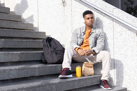 African american student sitting on campus steps listening to music with backpack and juice