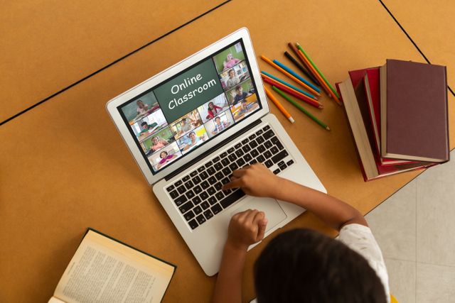 Student participating in online classroom lesson on laptop, surrounded by books and colored pencils. Perfect for use in articles and resources about e-learning, distance education, homeschooling, and remote classroom technologies. Can be used for educational websites, blogs about modern teaching methods, or promotional materials for online learning platforms.