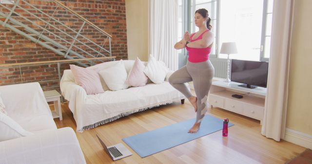 Woman is practicing yoga on a blue mat in a cozy living room with a laptop and water bottle nearby. Large windows and natural light fill the space, and a modern brick wall forms the backdrop. Ideal for content promoting home fitness programs, yoga tutorials, self-care routines, online yoga classes, and healthy living tips.