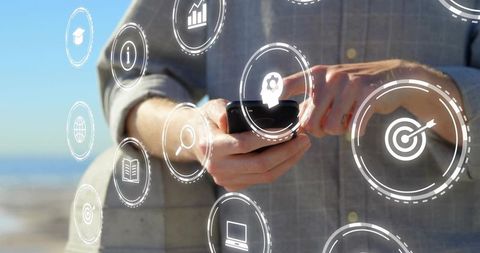 Holding smartphone and tapping digital interface with business icons on coastal background