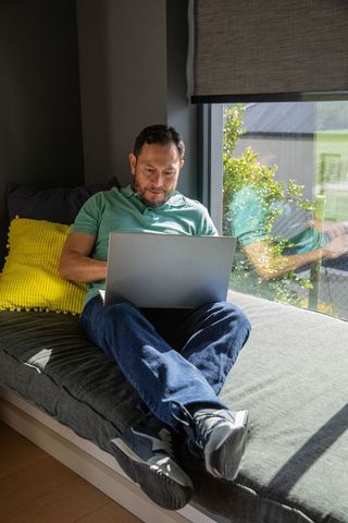 Mature asian man working on laptop in sunny window nook, relaxed modern home office