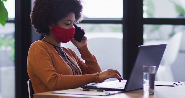 African-American woman wearing face mask working on laptop in office while talking on phone - Download Free Stock Images Pikwizard.com