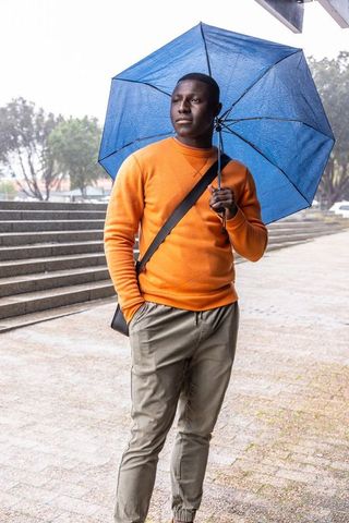 African american man standing under blue umbrella wearing orange sweater rainy urban plaza