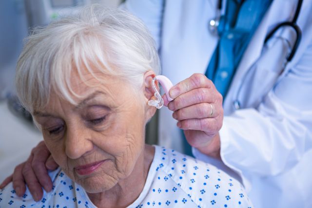 Doctor fitting hearing aid for senior patient in hospital. Useful for healthcare, medical treatment, elderly care, audiology, and hearing loss support concepts.