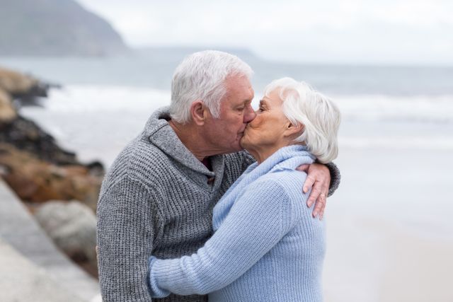 Senior couple sharing a romantic kiss on the beach, both wearing cozy sweaters. Ideal for use in advertisements, articles, or campaigns focused on love, romance, elderly lifestyle, retirement, and outdoor activities.