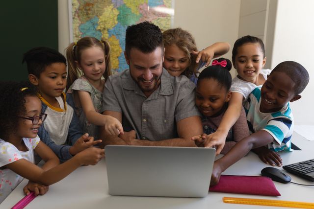 Male teacher engaging with diverse group of elementary school students around a laptop in a classroom. Children are actively participating and pointing at the screen, showing enthusiasm and collaboration. Ideal for use in educational materials, technology in education promotions, and diversity in learning environments.