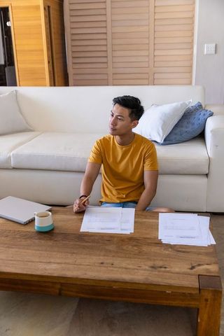 Contemplating Asian man reviewing documents on living room floor with coffee