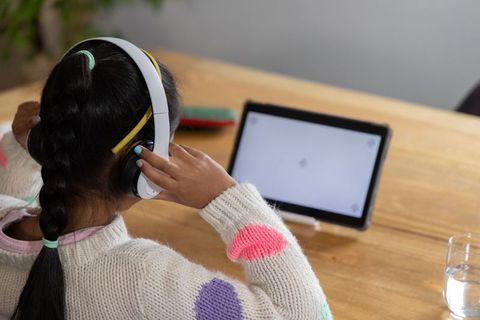 Child wearing headphones and sweater watching tablet at wooden table for remote learning