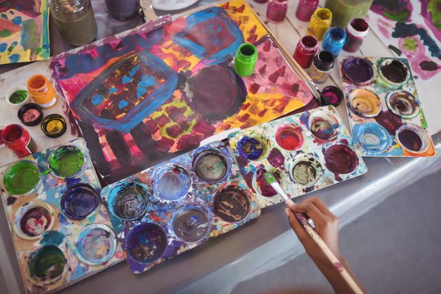This image shows a child's hand holding a paintbrush while working on a colorful painting. The desk is filled with various color palettes and paint jars, indicating a creative and artistic environment. This image can be used for educational content, art class promotions, children's creativity workshops, or any content related to childhood hobbies and artistic expression.