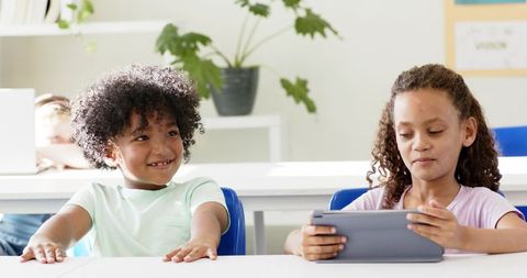 Multicultural elementary students sharing tablet, smiling and learning in bright classroom