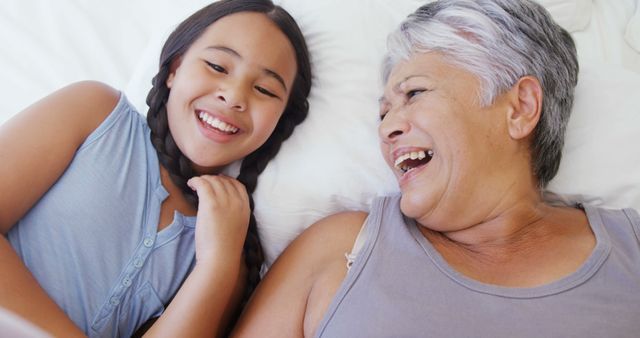 A young Asian girl enjoys a cheerful moment with her senior grandmother, lying together on a bed. Their laughter and close bond are evident as they share a joyful and intimate family time.