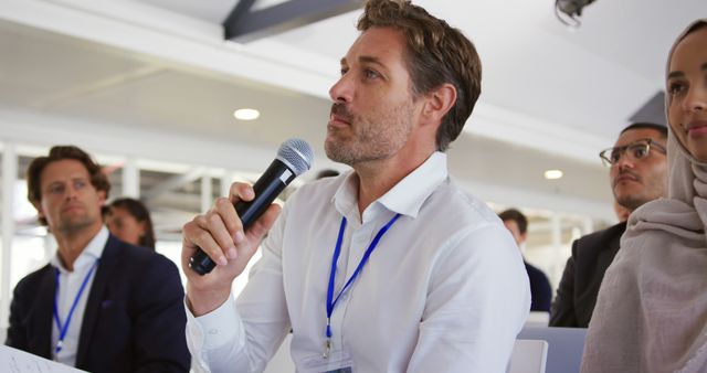 Middle-aged man holding a microphone, asking a question during a conference or seminar. Ideal for use in business-related articles, educational content, networking event promotions, or professional training materials.