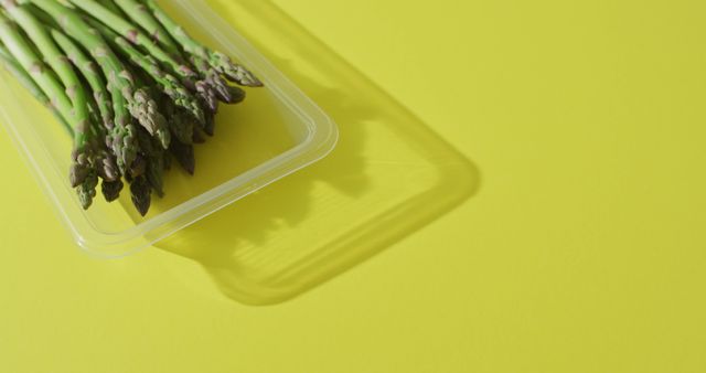 Bright image featuring a bunch of fresh asparagus in a clear plastic container, highlighted against a vibrant yellow background. Ideal for use in healthy eating campaigns, organic food promotions, and vegetarian or vegan cooking blogs. Visually appealing for food packaging designs or marketing materials focusing on fresh produce.