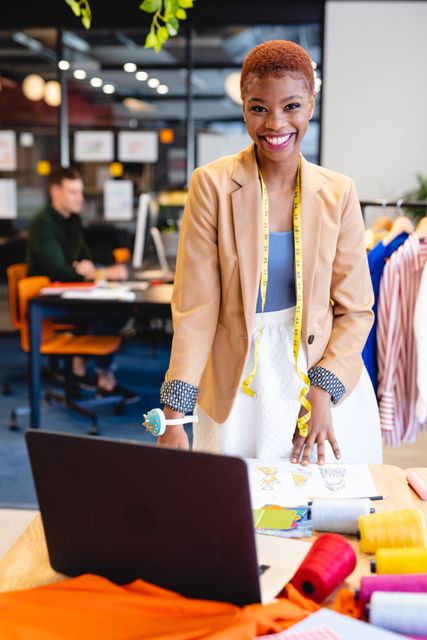 African American businesswoman in a modern office environment, enthusiastically engaging with her laptop. She exudes confidence and professionalism, possibly working in a creative or fashion-related field, as indicated by the measuring tape and rolls of fabric on the table. The cheerful expression and stylish clothing convey a positive and energetic work atmosphere. Perfect for articles or promotional materials on entrepreneurship, modern workspaces, professional women, or the fashion industry.