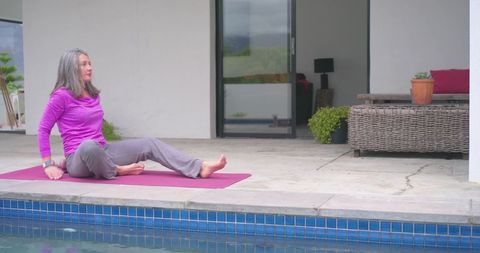 Mature woman practicing gentle yoga and stretching on magenta mat beside backyard pool