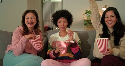 Friends night in: diverse women laughing, watching movie, eating popcorn on cozy couch