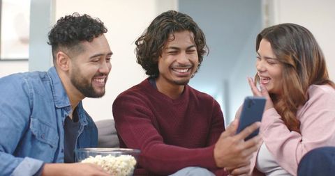 Friends laughing while sharing smartphone and popcorn on cozy living room couch