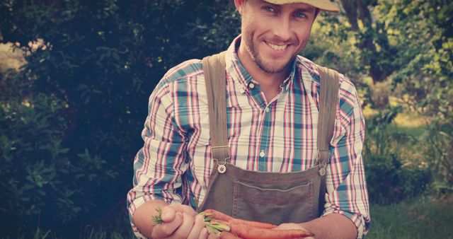 Smiling farmer holding freshly harvested carrots while wearing plaid shirt and overalls in garden. Ideal for content related to farming, gardening, organic produce, sustainable agriculture, farm-to-table movement, health and wellness.