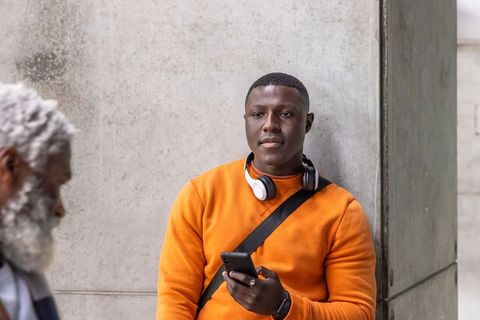 African american man leaning on concrete wall wearing orange top, headphones, smartphone