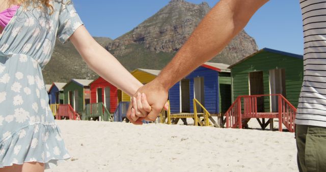 A young Caucasian couple holds hands on a sunny beach with colorful beach huts in the background, with copy space. Their casual attire and the romantic gesture suggest a leisurely vacation or a summer getaway.