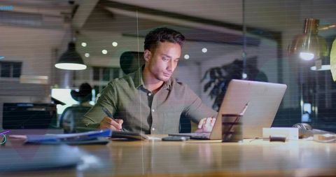 Hispanic professional working late on laptop in modern open-plan office taking notes