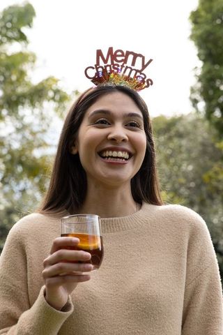 Asian woman smiling with merry christmas headband holding glass of amber drink in garden