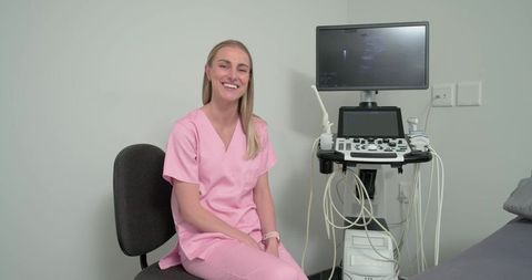 Smiling ultrasound technician wearing pink scrubs holding probe beside ultrasound machine
