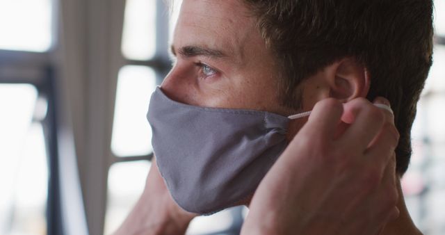 Person adjusting face mask with hands indoors, focusing on safety and health precautions. Ideal for articles or visuals related to health, personal safety, pandemic measures, and precautionary actions.