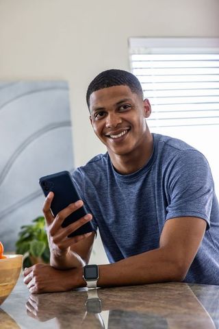 Smiling man leaning on counter checking smartphone wearing smartwatch displaying analytics