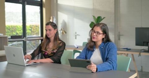 Diverse female colleagues collaborating with laptop and tablet in sunlit office meeting room