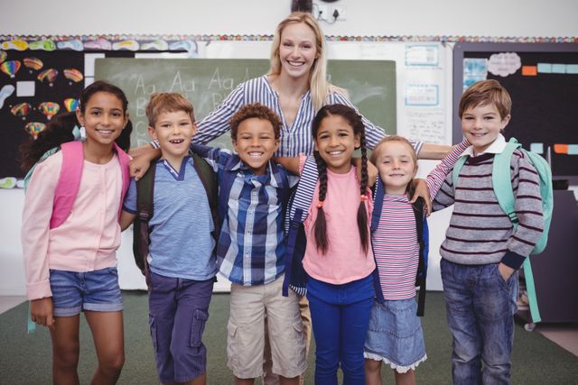 Teacher standing with a diverse group of smiling schoolchildren in a classroom. Children are wearing casual clothes and backpacks, indicating a school environment. This image can be used for educational materials, school advertisements, diversity and inclusion campaigns, and articles about primary education.