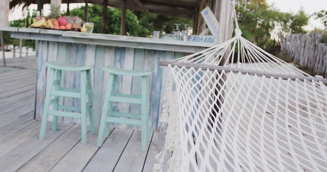 Hammock swaying in front of a rustic beach bar with wooden turquoise bar stools. Basket of tropical fruits and lush greenery in background. Perfect for depicting tropical vacation, summer getaway, relaxation, and leisure activities imagery.