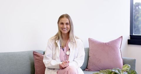 Smiling female clinician in pink scrubs and white coat on modern sofa in clinic office