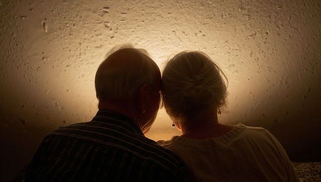 Senior couple sitting close on sofa, warm lamp halo creating intimate backlit silhouette