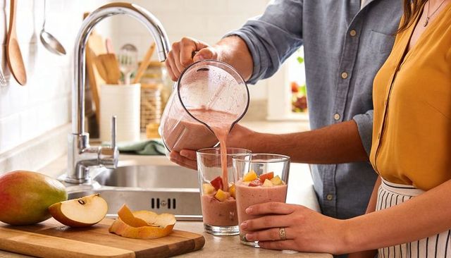 Couple pouring pink fruit smoothie from blender into glasses on sunlit kitchen counter