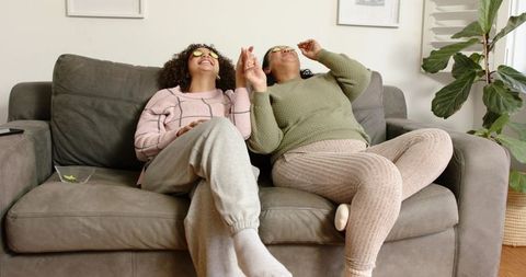 Two women relaxing on sofa with citrus eye pads, cozy sweaters at-home self-care