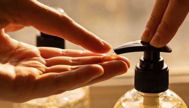 Hands pressing soap dispenser pump dispensing amber droplet backlit warm morning light